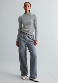 Vrouw met een aansluitende grijze gestreepte top met lange mouwen, losse grijze trainingsbroek met witte strook en witte loafers, staand tegen een effen achtergrond.