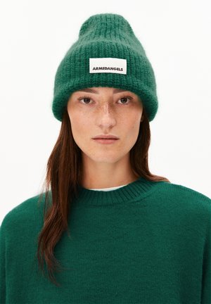 Green knitted beanie with a textured finish, featuring a white label with black text. Worn with a matching green sweater.