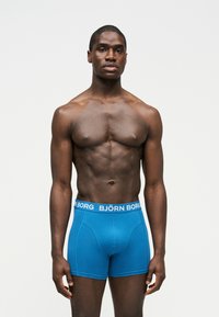 Homme musclé debout face à nous, portant un boxer bleu Björn Borg sur un fond blanc uni.