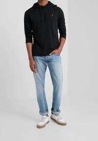 Svart hoodie med känguruficka och röd logotyp, i kombination med ljusblå denimjeans och vita sneakers med en beige sula.