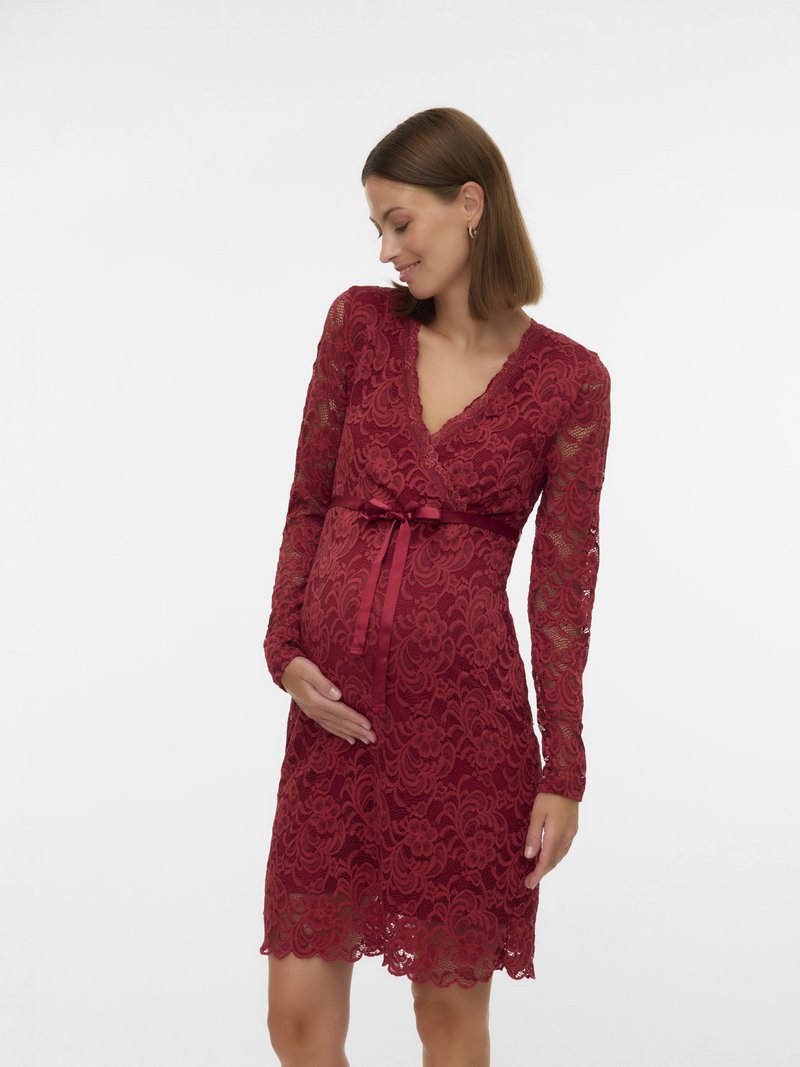 Έγκυος γυναίκα με μακρυμάνικο κόκκινο δαντελωτό φόρεμα και σατέν ζώνη, κρατώντας απαλά την κοιλιά της και κοιτώντας κάτω με χαμόγελο.