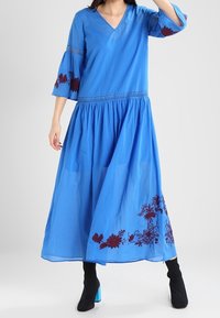 Blauwe lange jurk met een V-hals, uitlopende mouwen en bordeauxrode bloemenborduring nabij de zoom. Gemaakt van lichtgewicht stof, met een vloeiende uitstraling.