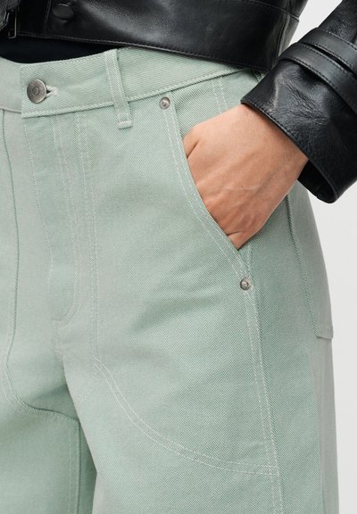 Άτομο που φοράει ανοιχτό πράσινο τζιν με καρφιά και μπροστινές τσέπες, με το χέρι στην τσέπη, σε συνδυασμό με μανίκι μαύρου δερμάτινου μπουφάν ορατό.