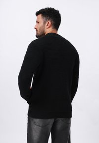 Pull en maille noir avec un motif texturé, col rond et manches longues. La vue de dos montre une coupe décontractée et un ourlet côtelé.