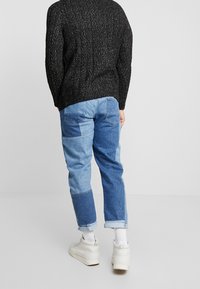 Pull en maille noire avec une finition texturée, associé à un jean en denim patchwork bleu avec des nuances variées et des revers roulés. Baskets blanches visibles.