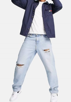 Personne portant un jean déchiré bleu clair, des baskets blanches, un t-shirt blanc et une chemise bleu marine à boutons avec un logo sur la poche poitrine.