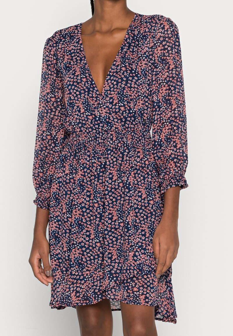 Femme portant une robe bleu marine avec un petit imprimé floral rose et blanc, manches longues, col en V et taille froncée.