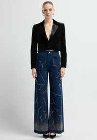 Blazer corto de terciopelo negro con tachuelas brillantes y solapa de satén, combinado con vaqueros de mezclilla azul oscuro de talle alto con patrones metálicos.