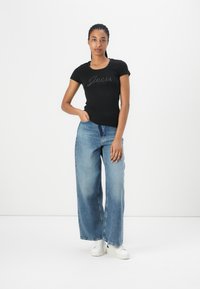 Black short-sleeve t-shirt with "Guess" logo in rhinestones, paired with loose light blue denim jeans and white sneakers.