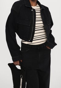 Chaqueta de mezclilla negra recortada con bolsillos frontales, combinada con una blusa a rayas en negro y crema, y pantalones de mezclilla negros. Sosteniendo un bolso negro texturizado.
