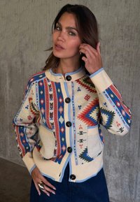 Γυναίκα που διορθώνει τα μαλλιά της, φορώντας πολύχρωμη ζακέτα με μεγάλα κουμπιά, που στέκεται μπροστά σε υφή τσιμεντένιου τοίχου.