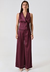Completo in satin bordeaux con gilet smanicato doppiopetto e pantaloni a gamba larga. Texture liscia, design minimalista e senza motivi visibili.