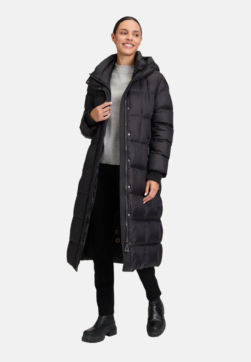 Schwarzer langer Puffermantel mit Kapuze, ausgestattet mit einem Reißverschluss vorne, gerippten Bündchen und einem steppierten Muster. Schwarze klobige Stiefeletten runden den Look ab.