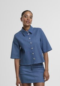 Cămașă din denim, scurtă, cu mâneci scurte, de culoare albastru deschis, cu închidere prin nasturi și un finisaj curat. Asortată cu o fustă din denim de aceeași nuanță.