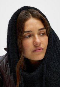 Svart, strukturerad stickad scarf som är virad runt huvudet och halsen, med ett mjukt, varmt utseende. Slät hy och subtil läppfärg syns.