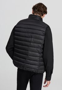 Gilet imbottito nero trapuntato con colletto alto, caratterizzato da una texture liscia e cuciture orizzontali. Indossato sopra una maglietta nera a maniche lunghe.