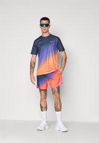 Herre atletisk outfit med en gradient T-shirt, der går fra marineblå til orange, og matchende shorts med pink accenter. Hvide sneakers fuldender looket.