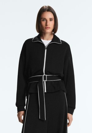 Žena s rovnými hnedými vlasmi nosí čiernu bundu na zips s bielym lemovaním, opásanú v páse, a zladené čierne nohavice s bielymi pruhmi.