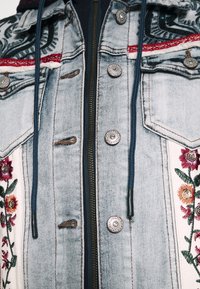 Veste en denim avec broderie florale à l'avant, dotée de boutons en argent, d'une fermeture éclair, de cordons de serrage bleu foncé et d'un délavage bleu clair.