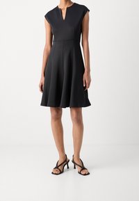 Robe noire sans manches avec un col en V, une jupe évasée et une texture lisse. Portée avec des sandales à talons hauts noires à lanières.