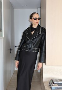 Must musta lõikekeelne jakk kontrastsete valgete õmblustega, vöö ja ülipaksu kraega, kombineeritud siresti musta kleidiga.