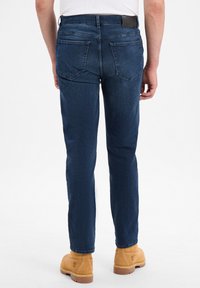 Ciemnoniebieskie dżinsy o dopasowanym kroju, z dwiema tylnymi kieszeniami i teksturowaną czarną skórzaną metką przy pasku. Widoczne brązowe botki.