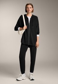 Felpa nera con cappuccio e zip, dotata di tasche frontali, abbinata a pantaloni sudati nere a gamba affusolata. Il modello indossa sneakers bianche e tiene una borsa chiara.