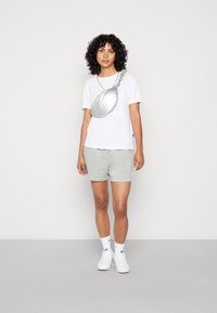 T-shirt blanc, shorts gris, sac en bandoulière argenté avec chaîne, baskets blanches et chaussettes blanches à mi-mollet. Cheveux bouclés. Fond neutre.