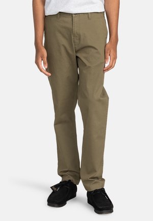 Olive green chinos with a regular fit, featuring a classic zipper and button closure, and side and back pockets. Worn with black shoes.