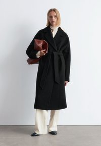 Schwarzer Wollumlage-Mantel mit großen Revers und taillierten Bund, kombiniert mit cremefarbenen Hosen und einer braunen strukturierten Handtasche. Flache schwarze Schuhe.