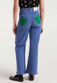 Persona con pantalones vaqueros azules y parches de corazón verdes en los bolsillos traseros, vistiendo mocasines negros brillantes, de pie frente a un fondo liso.