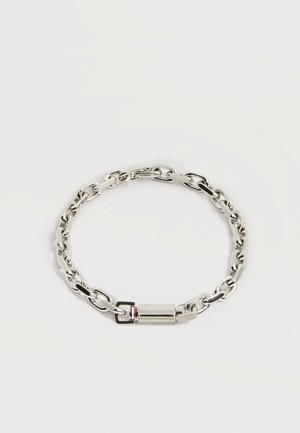 Srebrna bransoletka łańcuszkowa z owalnymi ogniwami i cylindrycznym zapięciem z małym czerwono-biało-czarnym detalem na jasnym tle.