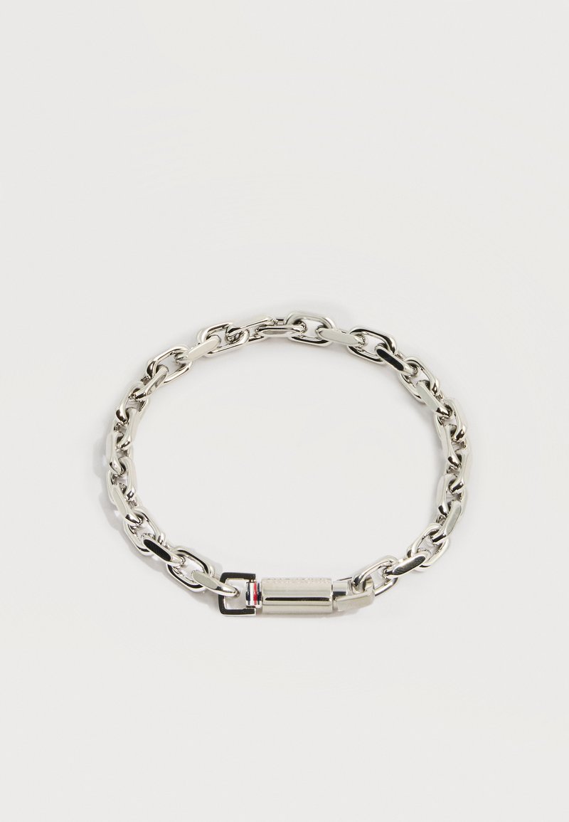 Bracelet en chaîne argentée avec des maillons ovales et un fermoir cylindrique présentant un petit détail rouge, blanc et noir sur un fond clair.