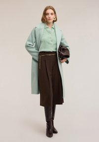 Manteau vert menthe texturé sur un haut vert clair, associé à un pantalon marron avec un détail de chaîne dorée et des bottes marron montantes jusqu'aux genoux.