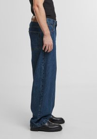 Donkerblauwe denim jeans met een losse pasvorm, rechte pijpen, twee achterzakken en een leren riem met een merkembleem, gecombineerd met zwarte schoenen.