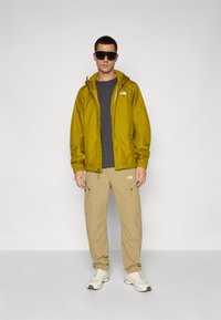 Veste jaune résistant à l'eau, chemise grise, pantalon beige et baskets blanches. Lunettes de soleil et accents logo sur les vêtements. Texture lisse.