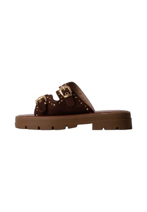 Sandale mule en daim marron avec semelle épaisse en caoutchouc, deux boucles dorées réglables et petits clous dorés le long des sangles.