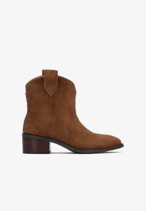 Ruskea mokkanahkainen nilkkatossu, jossa on pinottu puukorko, terävä pää ja vetolenkit sivussa.