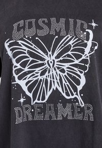 Černé tričko s grafickým motivem stříbrného motýla se skvrnami, zdobené akcenty z kamínků se nápisem "COSMIC DREAMER."