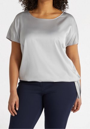 Femme portant un chemisier en satin argenté à manches courtes, rentré dans un pantalon ajusté bleu marine, debout avec les bras détendus le long du corps.