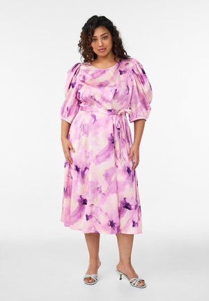 Žena v midi šatách s akvarelovým vzorom v ružovej a fialovej farbe, s nadýchanými rukávmi a viazaním v páse, doplnených bielymi sandálmi na podpätku.