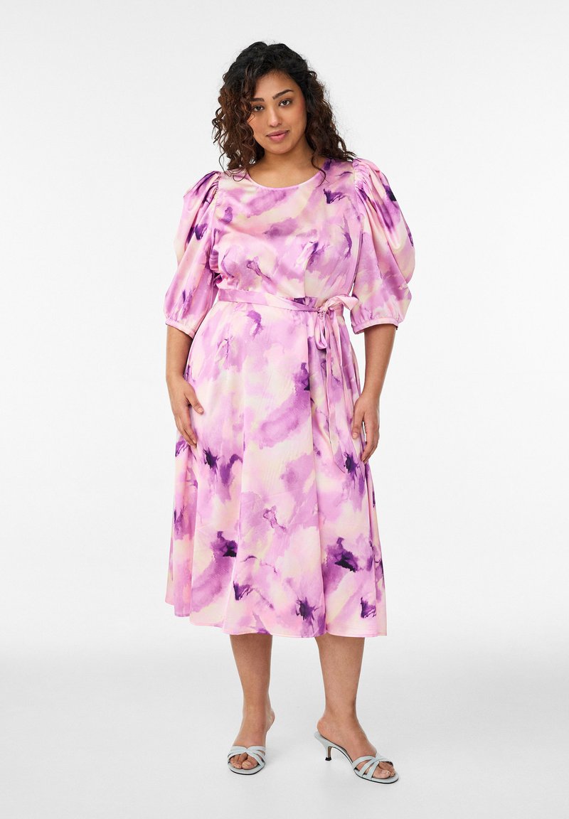 Frau trägt ein midi Kleid mit Aquarellmuster in Rosa und Lila, Puffärmeln und gebundener Taille, kombiniert mit weißen Sandalen mit Absatz.