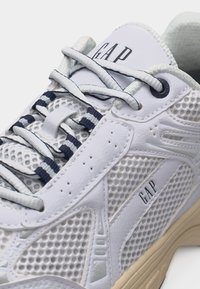 Baskets blanches de sport avec panneaux en mesh, lacets rayés blanc et marine, logo "GAP" sur la languette et le côté, semelle beige.