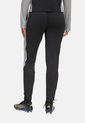 Osoba ubrana w czarne spodnie sportowe z białymi paskami po bokach i czarne korki z neonowożółtymi akcentami, stojąca tyłem.