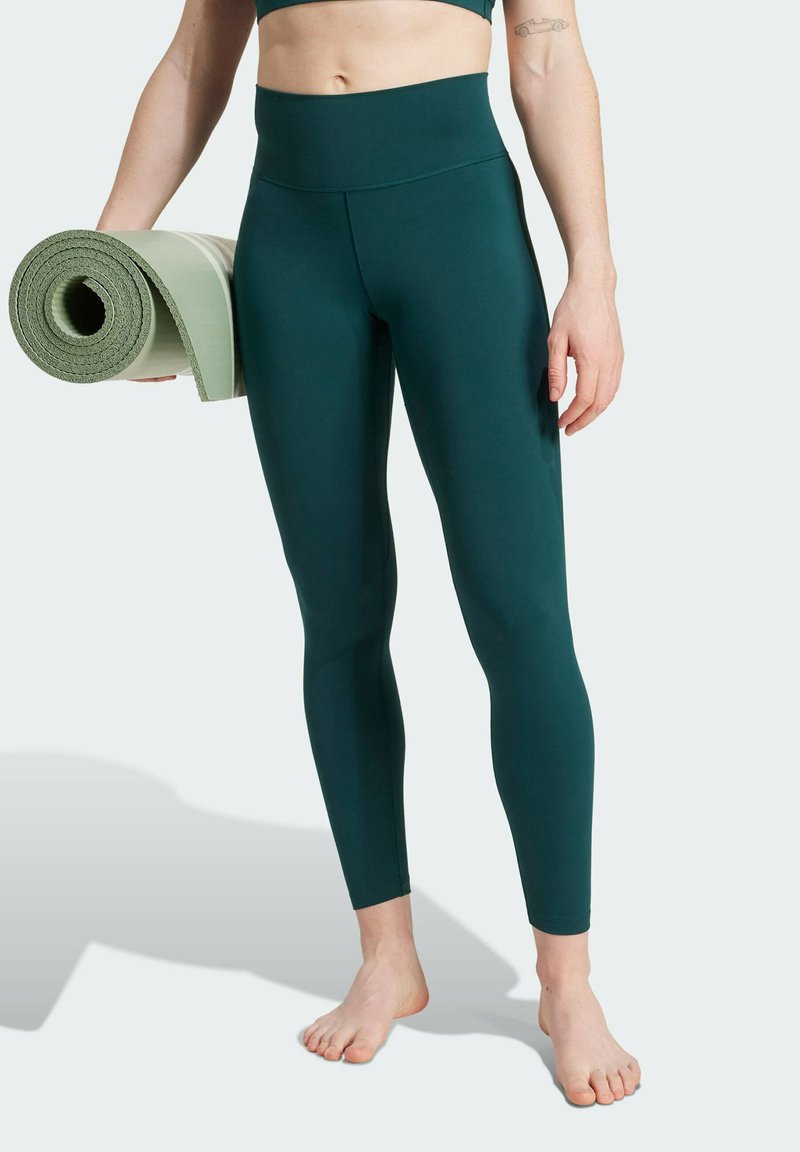 Leggings verde scuro realizzati in tessuto elastico, con una cintura alta. Un tappetino da fitness arrotolato di colore verde chiaro è tenuto in una mano.