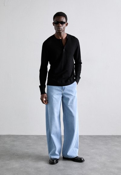 Zwarte langemouwen henley-shirt met een knoopsluiting en geribbelde textuur, gecombineerd met lichtblauwe, wijde jeans en glanzende zwarte schoenen.