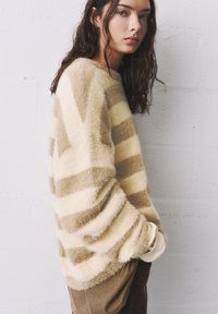 Beige-raita neule, joka on valmistettu pehmeästä ja karheasta materiaalista. Siinä on leveät hihat, rento malli ja pyöreä pääntie, joka on yhdistetty raidallisiin ruskeisiin housuihin.