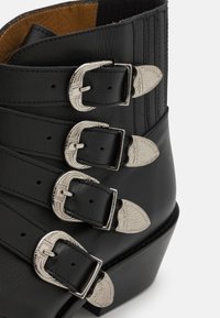 Botină din piele neagră până la gleznă, cu patru catarame argintii și accente decorative, având un vârf ușor conic și un panou superior texturat.