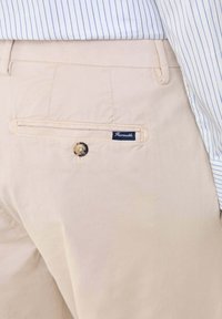 Pantaloni di cotone beige con una tasca posteriore dotata di un'etichetta blu e di un bottone rotondo. Il tessuto ha una consistenza liscia e una leggera lucentezza.
