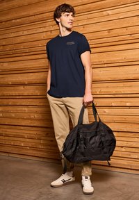 Svart duffelväska med justerbara remmar, texturerat tyg och flera fickor. Modellen bär en marinblå t-shirt och khakifärgade byxor, tillsammans med vita sneakers.